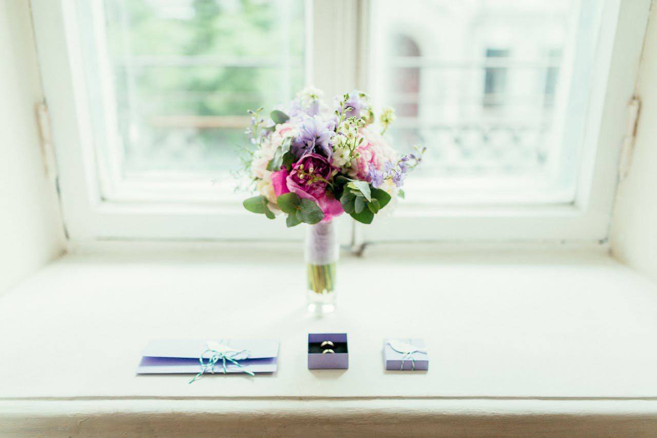 An elegant floral bouquet with wedding rings and gifts on a windowsill, captured in soft lighting.